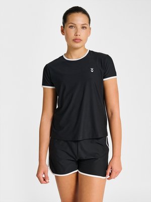 Kvinde står iført sorte atletikshorts og matchende sort t-shirt med hvid kant på ærmer, krave og shorts' kanter.