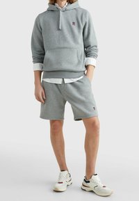 Felpa grigia con tasca anteriore e un piccolo logo. Abbinata a pantaloni corti grigi e sneakers bianche. Il materiale sembra morbido e confortevole.