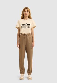 T-shirt en coton beige avec un graphique "Cool Club", associé à un pantalon à cordon de serrage marron avec des poches avant, le tout porté avec des sneakers beiges.