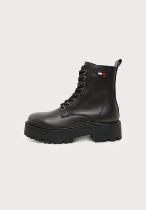 Botín de cuero negro con cordones, suela gruesa con tacos y pequeño parche rectangular con logo cerca de la parte superior en el lateral exterior.