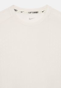 T-shirt Nike beige clair avec un col rond, présentant un motif texturé et un logo noir subtil au niveau du col. Tissu doux et lisse.