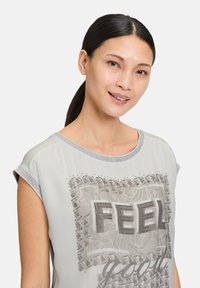 Chemise à manches courtes gris clair avec un design superposé, un fond floral, des accents cloutés, et le texte "FEEL good" en lettres métalliques.