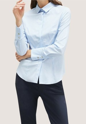 Kvinde iført lyseblå skjorte med striber og lange ærmer samt mørke bukser, armene krydsede og den ene hånd løftet nær ansigtet.