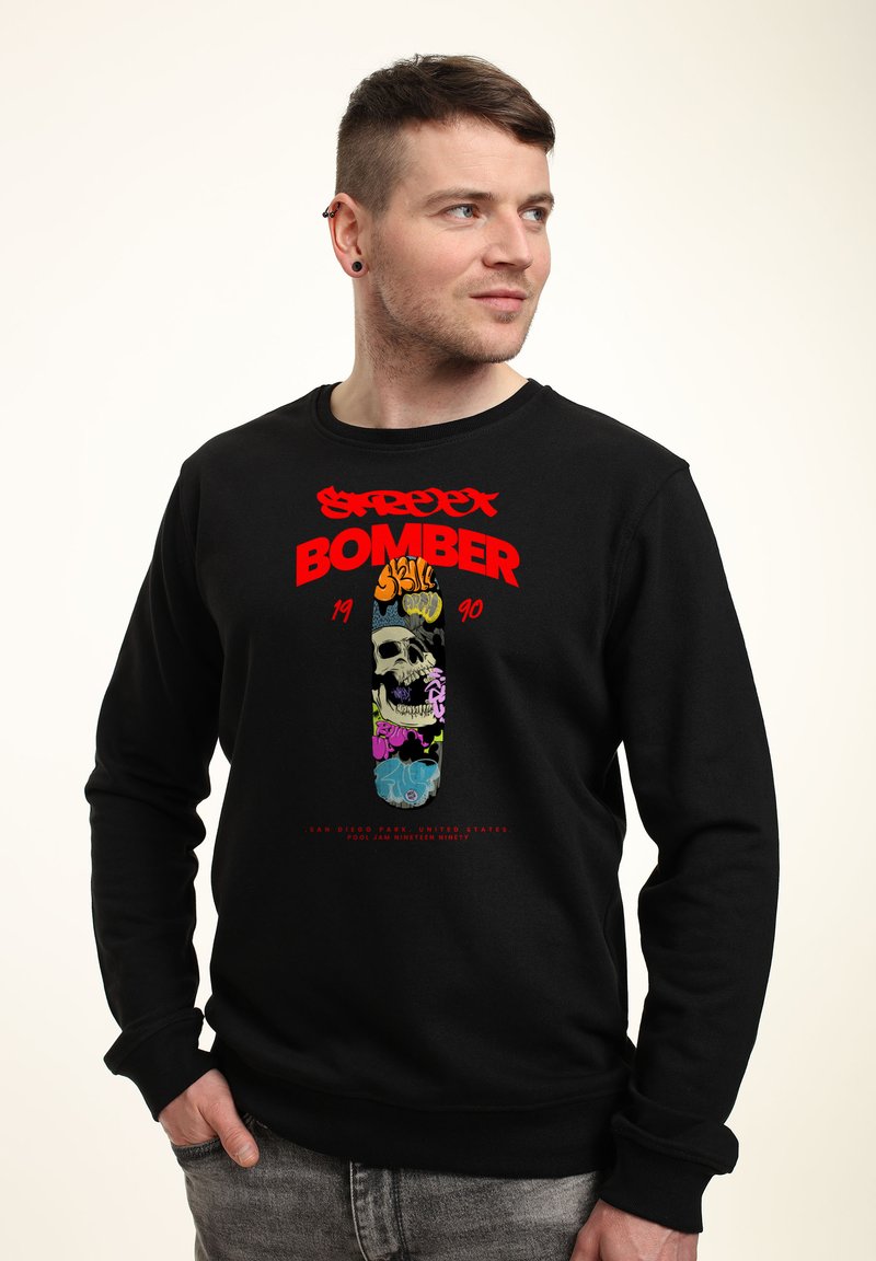Svart sweatshirt med en färgglad grafisk design av ett dödskalle med graffiti-inslag, fet röd text "STREET BOMBER" och ribbad mudd.