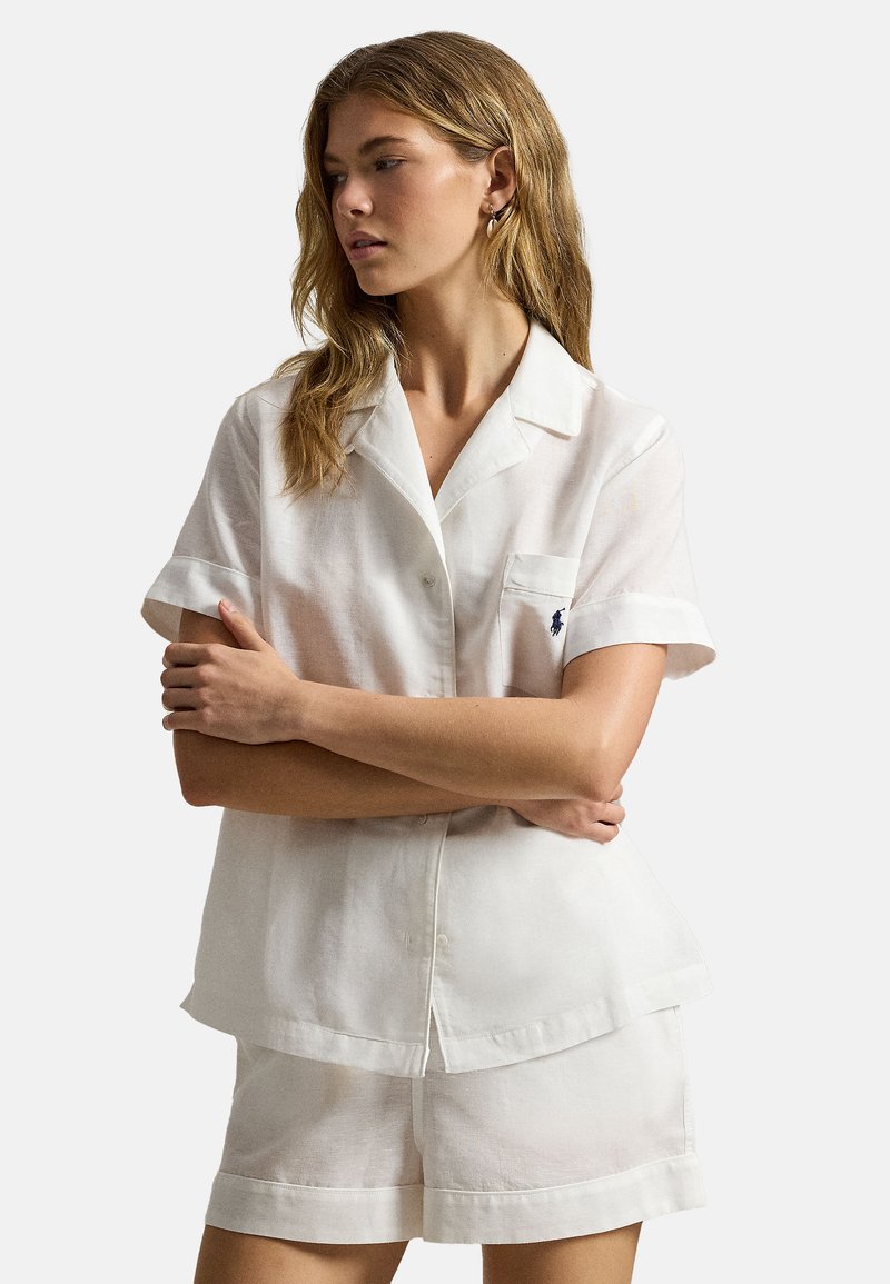 Jonge vrouw met lang lichtbruin haar, gekleed in een witte korte-mouwen blouse met knopen en bijpassende korte broek, armen gekruist, kijkend naar de zijkant.