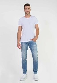 Camiseta blanca de cuello en V para hombre, combinada con jeans ajustados de color azul claro y zapatillas blancas. La tela parece suave con un diseño minimalista.