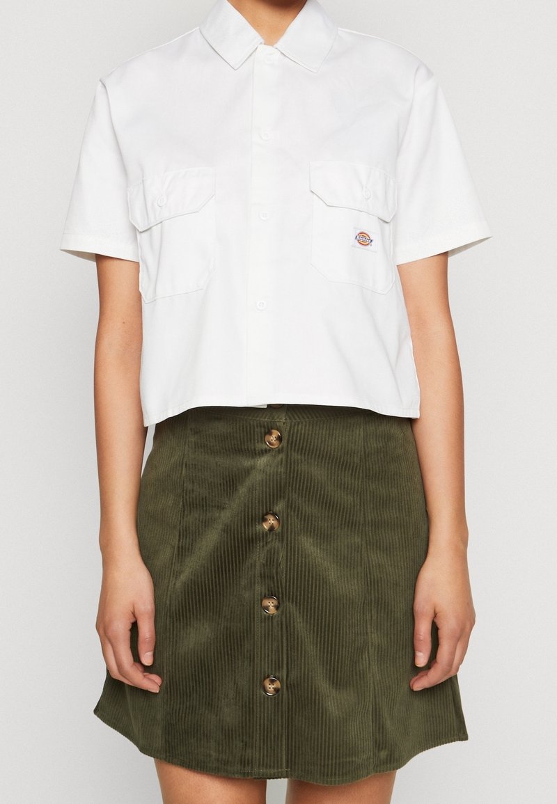 Chemise blanche à manches courtes avec boutons, deux poches avant et logo, associée à une jupe en velours côtelé vert olive boutonnée devant.