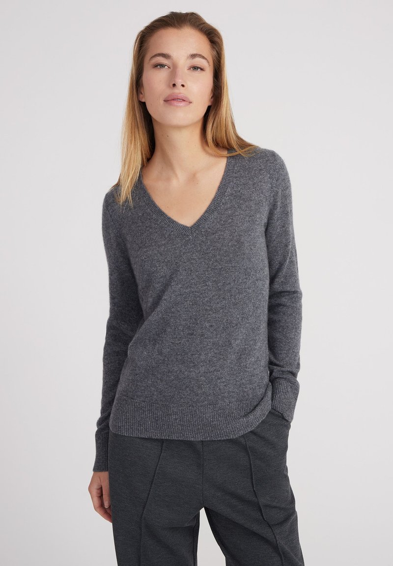 Grauer V-Ausschnitt-Pullover aus weichem Strick mit langen Ärmeln und geripptem Saum. Über einer dunklen Hose getragen, mit lässiger Passform und glatter Textur.