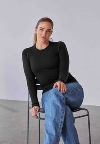 Chemise noire à manches longues avec un col rond, associée à un jean en denim bleu clair. Le tissu semble lisse avec une silhouette ajustée.