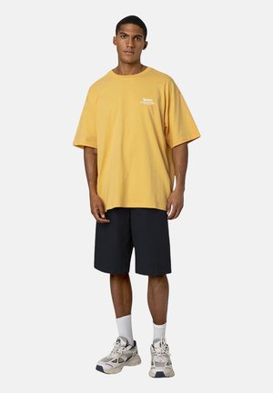 Gelbes, übergroßes T-Shirt aus Baumwolle mit kurzen Ärmeln, das weißen Text auf der Brust trägt. Kombiniert mit schwarzen Shorts und beigen Sneakers.