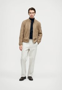 Καφέ σουέτ bomber jacket με ψηλό γιακά, συνδυασμένο με μπλέ τζέρσεϊ και ανοιχτού γκρι παντελόνι, ολοκληρωμένο με σκούρα παπούτσια.