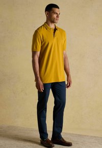 Kollane polo särk, millel on krae ja lühikesed varrukad, kombineerituna tumesiniste pükstega ja pruunide loafersitega, seistes neutraalses taustas.