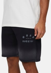 Pantaloni corti neri con una texture liscia, caratterizzati da una grafica stampata "SAND for NOTHING" in grigio sul lato sinistro.