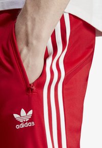 Rote Sportshorts mit einem Reißverschlussfach, drei weißen Streifen an der Seite und einem bestickten Adidas-Logo. Glatte Textur.