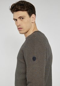 Sweter z dzianiny z teksturowanym, geometrycznym wzorem w kolorze brązowym i granatowym, z czarną naszywką logotypu na rękawie. Okrągły dekolt.