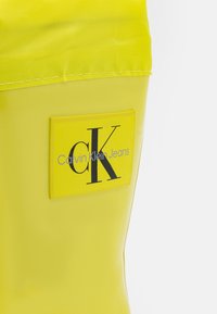 Bolsa impermeable amarilla con una textura suave, que presenta una etiqueta rectangular con "cK" y "Calvin Klein Jeans" en impresión negrita de color negro.