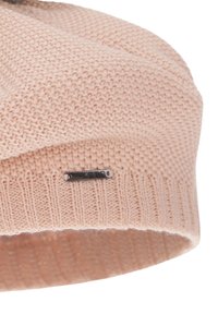 Gebreide beanie in lichtroze, met een geribde band en een klein metalen label. De textuur is zacht en iets los zittend.