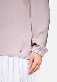 Light pink long-sleeve top with gathered cuffs, featuring a small brown accent detail at the hem. Smooth fabric texture.