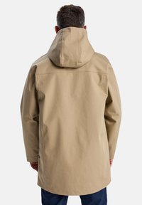 Une personne portant un imperméable beige avec une capuche est vue de dos, se tenant contre un fond blanc uni.