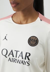 Fehér sportpóló világos rózsaszín kiegészítőkkel, fekete logóval a Paris Saint-Germain számára és vastag fekete betűkkel a "QATAR AIRWAYS" felirattal.