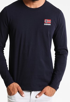 Man wearing a navy blue long-sleeve shirt with a small Norwegian flag logo on the upper left chest, hands in light-colored pants pockets.