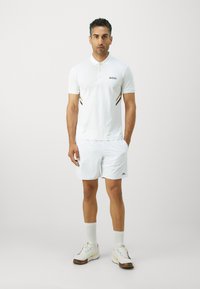 White polo shirt with BOSS logo, featuring vertical stripes in black and gold. Paired with white shorts and multi-colored athletic shoes.