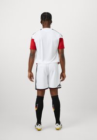 Λευκή και κόκκινη στολή ποδοσφαίρου με μαύρες λεπτομέρειες. Τιράντα κοντού μανικιού, σορτσάκι και μαύρες κάλτσες που περιλαμβάνουν λεπτομέρεια γερμανικής σημαίας.