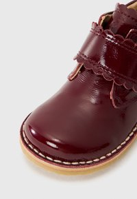 Sapato de couro patente cor burdeos com detalhe de tira recortada, bico arredondado e costura em contraste numa sola bege. Design compacto para crianças.