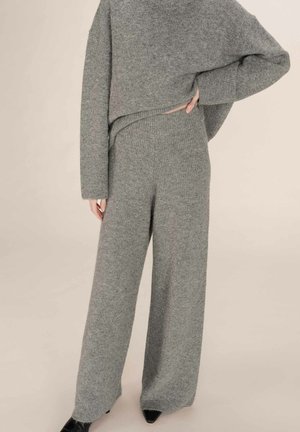 Pantalon large en tricot gris associé à un pull loose assorti, porté par une personne debout avec une main sur la hanche et des chaussures noires à bouts pointus visibles.