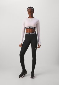 Top cropped à manches longues Nike Pro en blanc, associé à un legging noir taille haute avec une ceinture logo. Des chaussures de sport noires complètent la tenue.