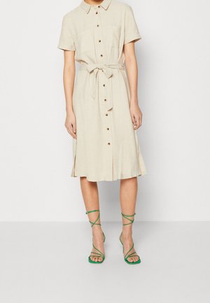 Femme portant une robe midi beige à boutons sur le devant, manches courtes et taille nouée, associée à des sandales à talons hauts vertes à lanières.