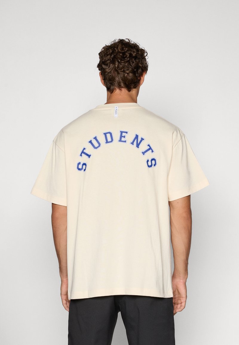 Beige bomull t-skjorte med korte ermer, med blå brodert "STUDENTS" i en buet design på baksiden. Ledig passform.