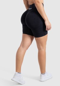 Zwarte naadloze shorts met een hoge tailleband, geribbeld textuur en gebogen naden voor verbetering. Gecombineerd met witte sportschoenen.