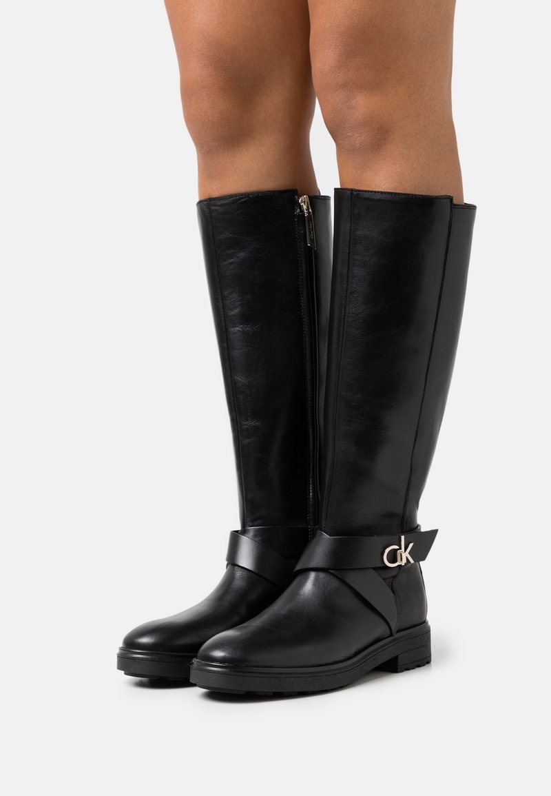 Bottes en cuir noir montant jusqu'au genou, avec talons bas, fermetures éclair latérales et sangles décoratives ornées d'un petit logo argenté "ck" sur la partie extérieure des chevilles.