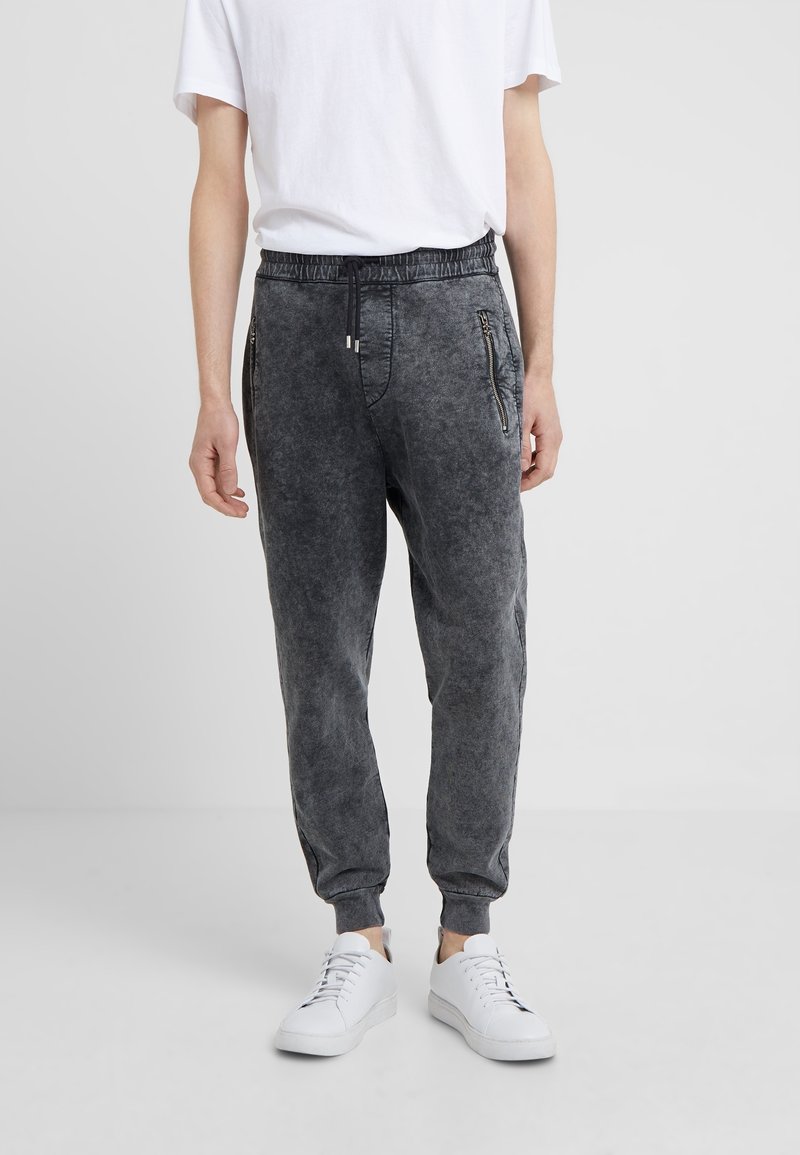 Person som bär tvättade svarta joggerbyxor med dragkedjor på sidorna, vita sneakers och en vanlig vit t-shirt mot en grå bakgrund.