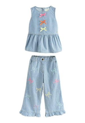 Mladé dievča má na sebe modro-biele pruhované tielko bez rukávov a nohavice s farebnými aplikáciami vo forme mašlí a zlaté sandále, stojí s miernym úsmevom.