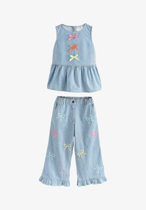 Fată tânără purtând un top fără mâneci cu dungi albastre și albe și pantaloni cu aplice colorate în formă de fundițe și sandale aurii, stând cu un zâmbet ușor.