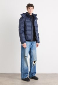 Navy puffer jacket with a zip front, hood, and large quilted sections, paired with light blue distressed wide-leg jeans and black shoes.