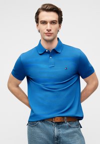 Polo bleu en tissu texturé avec une patte à trois boutons et un petit logo brodé. Porté avec un jean en denim clair et une ceinture marron.