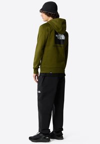 Felpa verde oliva con un grande logo nero sulla schiena, abbinata a pantaloni neri e scarpe nere. I materiali sembrano morbidi e resistenti.