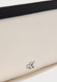 Κρεμ θήκη με υφή, με μαύρη φερμουάρ και μικρό ασημένιο λογότυπο "ck" στο μπροστινό μέρος.