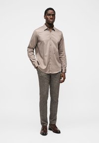 Ανοιχτό καφέ πουκάμισο με κουμπιά και μακριά μανίκια, σε συνδυασμό με υφασμάτινο γκρι παντελόνι και καφέ παπούτσια, φορεμένα από μια όρθια ανδρική φιγούρα.