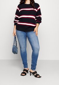 Strikket marineblå sweater med hvide og røde striber, parret med lyseblå jeans, sorte hæle-sandaler og en lille denim håndtaske.