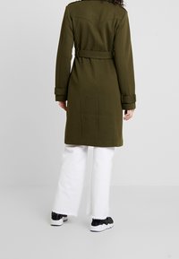 Olijfgroene jas met een gedefinieerde kraag, riemdetail en opgerolde mouwen, gecombineerd met witte wijde broek en zwarte sneakers.