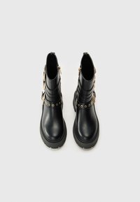 Bottes noires jusqu'à la cheville en cuir lisse, avec une semelle épaisse, des accessoires en métal doré décoratifs, et des fermetures éclair de chaque côté.