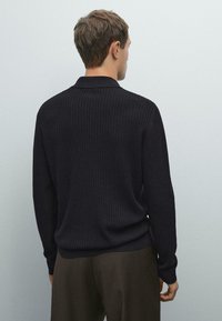 Man met zijn rug naar de camera, gekleed in een zwart geribbeld lange mouw trui en donkere broek, tegen een effen lichtgrijze achtergrond.