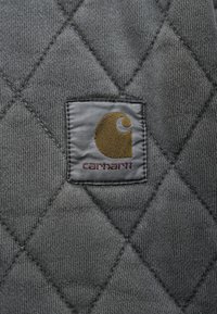 Tejido de quilt gris que presenta un parche rectangular gris con un logo circular dorado y el texto "carhartt" bordado en burdeos.