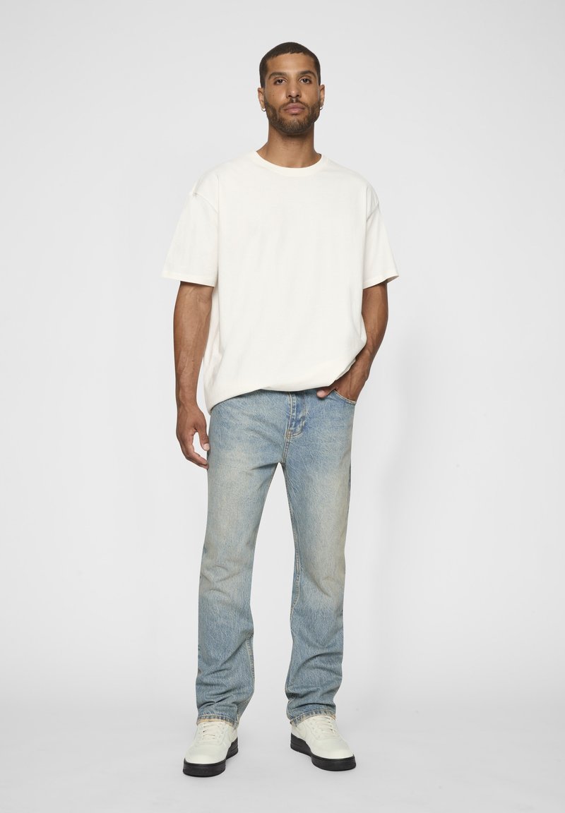 Man staat, draagt een eenvoudig wit oversized T-shirt, lichtblauwe spijkerbroek en witte sneakers met zwarte zolen, hand in zak.