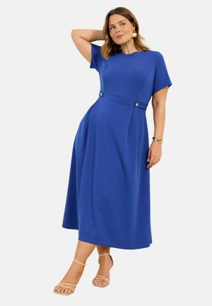 Frau in einem blauen Midikleid mit kurzen Ärmeln, die mit einer Hand hinter dem Kopf steht, beige Riemchensandalen und goldenen Schmuck trägt.