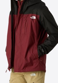 Homme portant une veste imperméable The North Face bordeaux et noire ainsi qu'un pantalon cargo beige avec logo, debout les mains dans les poches.